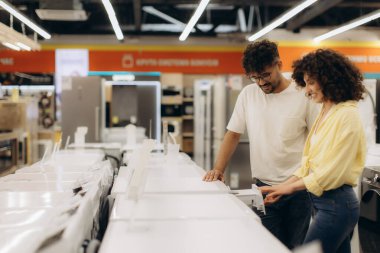 Mutlu bir çift bir elektronik mağazasını keşfediyor, beyaz çamaşır makinelerine ilgi ve merakla bakıyor. Parlak ışıklandırma mevcut aygıtların çeşitliliğini vurgular.