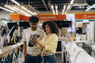 Bir çift, iyi ışıklandırılmış bir elektronik mağazasında alışveriş yaparken modern ütü çeşitlerini inceliyor. Mutlu ve meşgul görünüyorlar, birlikte alışveriş deneyimlerinden zevk alıyorlar..