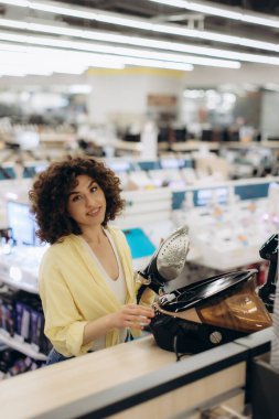 Modern bir elektronik mağazasındaki genç bir kadın buhar ütüsünün işlevselliğini kontrol ediyor. Ayar, yenilik, teknoloji ve alışveriş deneyimlerinin vurgulandığı çeşitli cihazlar içeriyor ve sergileniyor..