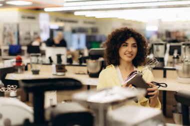 Neşeli bir kadın, parlak bir elektronik mağazasında çeşitli ürünleri sergileyen mutfak aletlerini inceliyor..