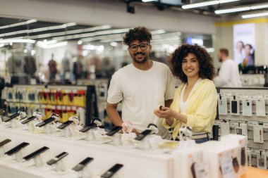 Bir elektronik mağazasının akıllı telefon bölümünde cihazları hevesle karşılaştıran neşeli bir çift. Parlak ortam, çeşitli elektronik aletler ve aksesuarlar için dostane ve modern bir alışveriş deneyimini geliştirir..