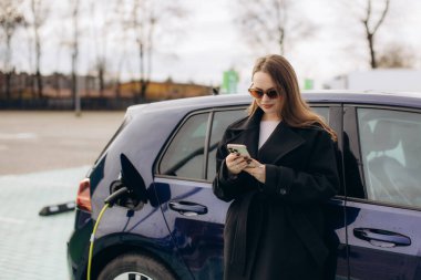 Elektrikli aracının yanında duran bir kadın şarj istasyonunda beklerken akıllı telefonunu kontrol ediyor.