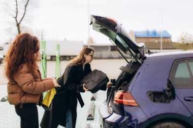 İki kadın, bir şarj istasyonunda mavi bir elektrikli arabanın bagajına eşya yerleştiriyor ve sürdürülebilir seyahat için hazırlanıyor.