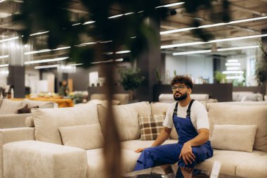 Modern bir mobilya mağazasında rahat bir kanepede dinlenen tulumlu bir adam molada etrafındakileri düşünüyor.