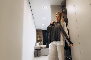 Genç bir kadın, elinde bir kazakla modern bir dolaba giriyor ve günlük kıyafetleri hakkında karar veriyor.