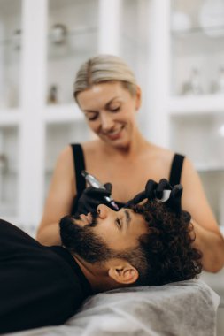 Kozmetik uzmanı erkek hasta, kaşlarına kalıcı makyaj uygularken güzellik salonunda yüz hatlarını geliştiriyor.