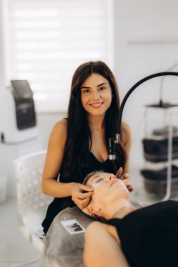 Güzellik uzmanı, modern güzellik salonundaki rahat bir bayan müşteriye doğal güzelliği geliştirmeye odaklanarak kirpik eklentileri uygularken gülümsüyor.