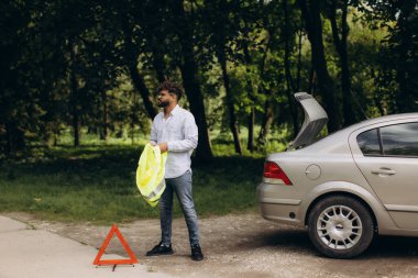 Uyarı üçgeninin yanında güvenlik yeleği tutan ve yol yardımını bekleyen bir adam var.