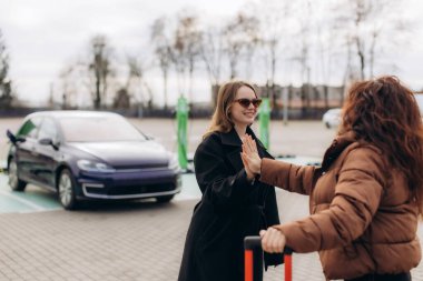 Kadın arkadaşlar buluşuyor, açık havada elektrikli araç şarj istasyonunda bir arabanın yanında bavulla beşlik çakıyorlar.