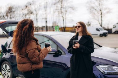 Arkadaşlar konuşuyor ve kahvenin tadını çıkarıyorlar. Elektrikli aracın açık hava istasyonunda şarj olmasını bekliyorlar.