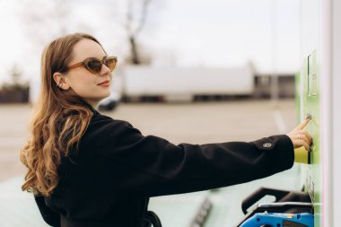 Kadın elektrikli arabası için halka açık bir şarj istasyonunu çalıştırıyor, çevre dostu enerjiyi ve modern yaşam tarzını sembolize ediyor.