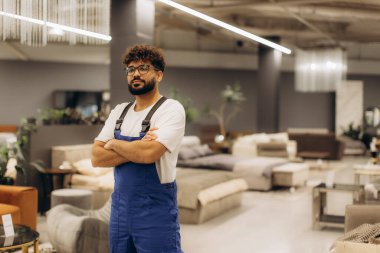 Tulumlu ve gözlüklü bir adam kollarını kavuşturmuş modern mobilya galerisini gururla denetliyor.