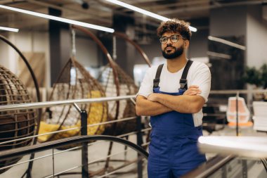 Tulum ve gözlük takan genç bir adam, kollarını kavuşturmuş, modern bir mobilya mağazasında güvenle duruyor.