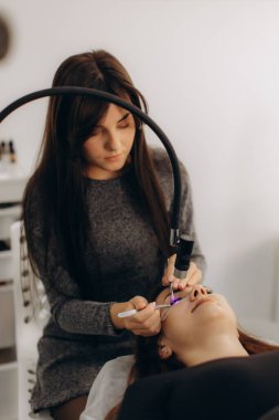 Kirpik uzatma uzmanı bir müşteriye cımbız ve ultraviyole iyileştirici lamba kullanarak özel kirpikleri dikkatlice uyguluyor.