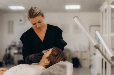 Profesyonel güzellik uzmanı, temiz bir salondaki güzellik prosedürü sırasında bir müşterinin kaşlarına kaş kaplaması tedavisi uyguluyor.