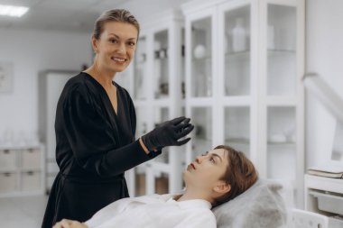 Siyah üniformalı ve eldivenli profesyonel güzellik uzmanı kuaförde yatan genç bir kadına kozmetik tedavi uygulamaya hazırlanıyor.