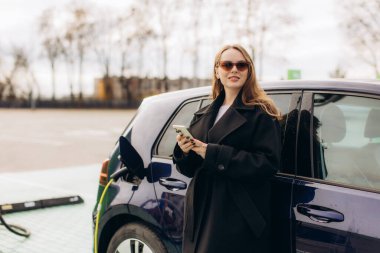 Elektrikli aracının yanında duran genç bir kadın, bir istasyonda şarj ederken akıllı telefon kullanıyor.