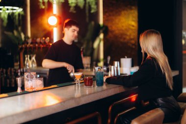 Zarif bir akşam ziyareti sırasında bir barmenin müşteriye renkli kokteyller hazırladığı şık ve çağdaş bir bar iç mimarisi. Çevre aydınlatması sıcak ve davetkar bir atmosfer yaratır..