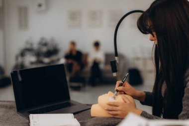 Genç bir kadın, dizüstü bilgisayarla güzellik dersi sırasında bir mankenin kafasına kirpik eklentisi uyguluyor.