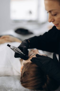 Siyah eldiven giyen kadın güzellik uzmanı bir güzellik salonunda müşterinin kaşlarına kalıcı makyaj tedavisi uyguluyor.