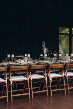 Ahşap sandalyeler, tabaklar, bardaklar ve çiçek aranjmanlarının yer aldığı bir şenlik ya da düğün için hazırlanmış açık hava uzun yemek masası.