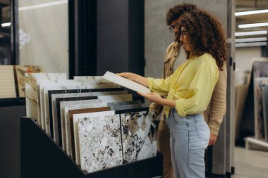 Genç çift yeni seramik fayanslar seçiyor. Kalıplar arasında seçim yapan bir ekranın yanında duruyorlar.