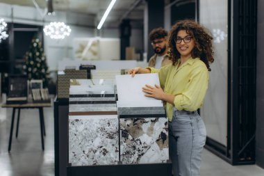 Fayans galerisinde elinde beyaz bir fayans örneğiyle duran ve gülümseyen bir kadın bir proje için yeni malzemeler seçerken...