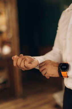 Adam hazırlanıyor, kollarını düzeltiyor. Beyaz resmi gömlek ve kolunda boncuklu bilezik olan turuncu akıllı saat.