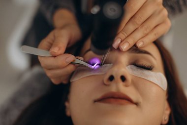 Cımbız kullanan bir güzellik uzmanından kirpik uzatma uygulaması alan bir kadın ve bağlanmak için özel UV ışığı