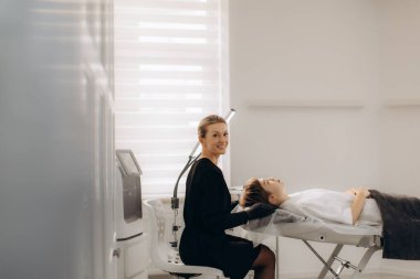Modern bir klinikte güzellik prosedürü için yatağa uzanmış bir müşteriyi hazırlarken kozmetolog gülümsüyor.