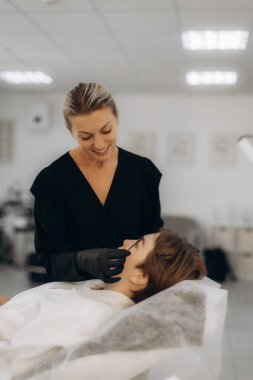 Siyah eldivenli profesyonel güzellik uzmanı güzellik salonundaki kozmetoloji kanepesinde dinlenen müşterisine kaş boyası uyguluyor.