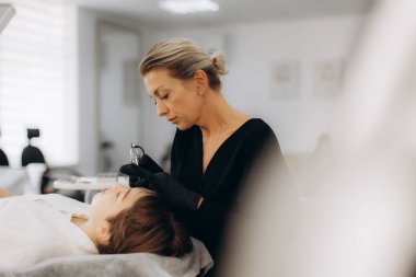 Modern bir güzellik salonunda genç bir kadının kaşlarına mikropla kalıcı makyaj yapan profesyonel bir güzellik uzmanı.