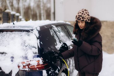 Karla kaplı bir elektrikli aracın yanında duran, şarj olan kabloyu takan ve soğuk bir kış gününde akıllı telefon kullanan bir kadın.