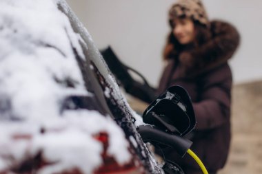 Karla kaplı elektrikli bir araca elektrik kablosu bağlayan kadın, sürdürülebilir enerji ve kış ulaşımını vurguluyor