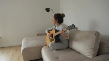 Kadın evde kanepede akustik gitar çalıyor. Kız müzik aleti çalıyor ve şarkı söylüyor. Yaratıcı hobi, boş zaman aktivitesi.