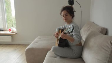 Kadın evde akustik gitar çalıyor. O, hobisinden hoşlanıyor ve oynarken gülümsüyor. Penceresi ve lambası olan rahat bir daire..