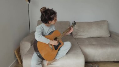 Kanepede oturan genç bir kadın akustik gitar çalıyor, evinde müziğin keyfini çıkarıyor. Boş zaman, müzik ve yaşam tarzı kavramları.