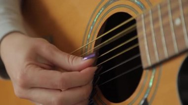 Akustik gitar çalan kadın ellerinin yakın çekimi. Kadın nazikçe telleri koparıp melodik bir ses çıkarıyor..