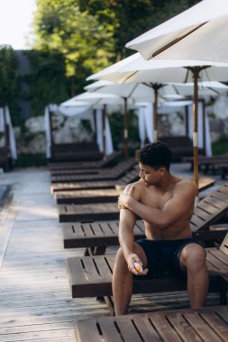 Genç adam havuz kenarında bir şezlongda oturuyor, omzuna güneş kremi sürüyor, güneşlenmeye ve dinlenmeye hazırlanıyor.