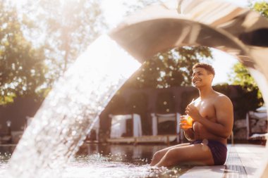 Genç adam tropikal bir otelde lüks bir yüzme havuzunda otururken ferahlatıcı bir kokteylin tadını çıkarıyor.