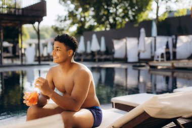 Genç adam havuz başında otururken renkli bir kokteylin tadını çıkarıyor. Lüks bir tatil köyünde, yaz havasını ve huzurlu atmosferi kucaklıyor.