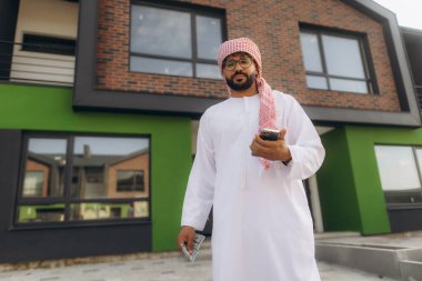 Parası ve akıllı telefonu olan bir Arap, zenginlik ve yatırımı simgeleyen, çağdaş yeşil ve tuğlalı bir evin önünde duruyor.