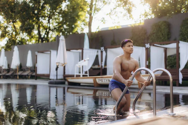 Genç atletik bir adam lüks bir otelde sakin bir yüzme havuzundan metal merdiveni kullanarak çıkıyor, ferahlatıcı bir yüzüşten sonra yaz güneşinin tadını çıkarıyor.