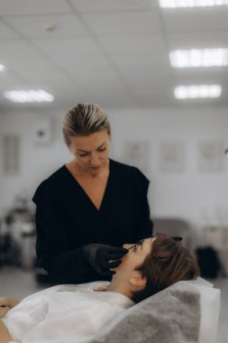 Siyah eldivenli bir güzellik uzmanı modern bir güzellik salonunda yatan kadın müşteriye kalıcı kaş makyajı yapıyor, profesyonel bakım ve hassasiyet.