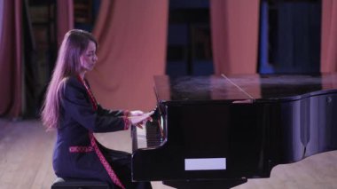 Sahnede piyano çalan genç bir kadın. O, kırmızı işlemeli koyu renk bir ceket giyiyor. Perdeler kapalı..