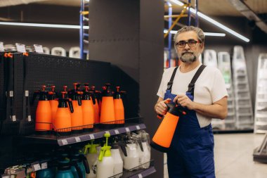 Tulum ve gözlük takan deneyimli bir işçi dükkan koridorunda turuncu bahçe spreyi tutuyor, ekipman alışverişi yapıyor.