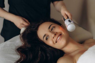 Mutlu kadın, profesyonel bir kozmetoloji kliniğinde cilt bakımı için gelişmiş bir cihaz kullanarak modern bir yüz tedavisi görüyor.