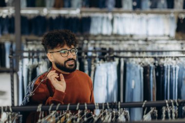 Sakallı ve gözlüklü genç şık bir adam modern bir butikte kot pantolon raflarını karıştırıyor.
