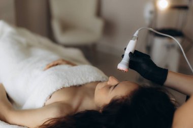 Yatağa uzanmış bir kadın, modern bir cihaz kullanarak bir profesyonelden yenilenme yüz bakımı alıyor.
