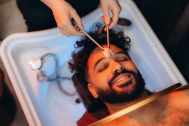 Sandalyede dinlenen bir adam, rahatlatıcı bir spa tedavisi görüyor, kafa sağlığı ve saç bakımına odaklanıyor.
